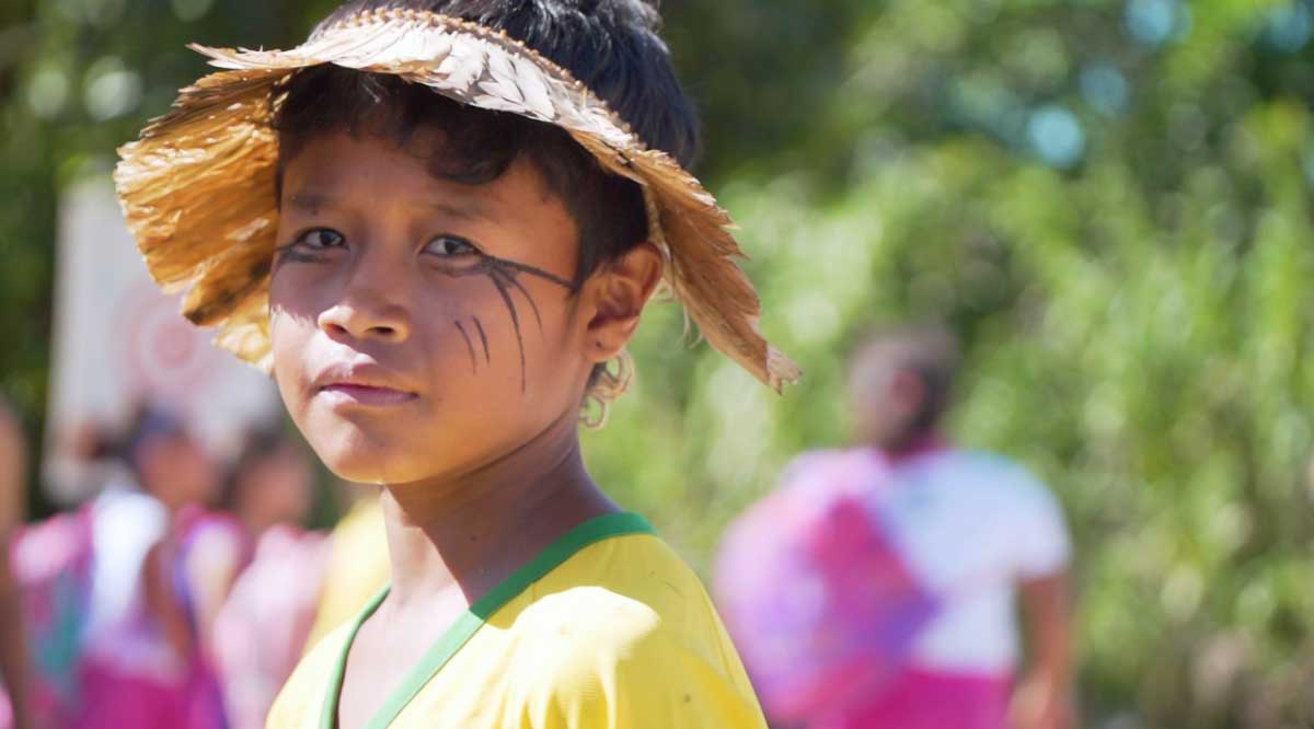 Menino indígena com o rosto pintado, um pequeno cocar e uma camiseta amarela com a gola verde.